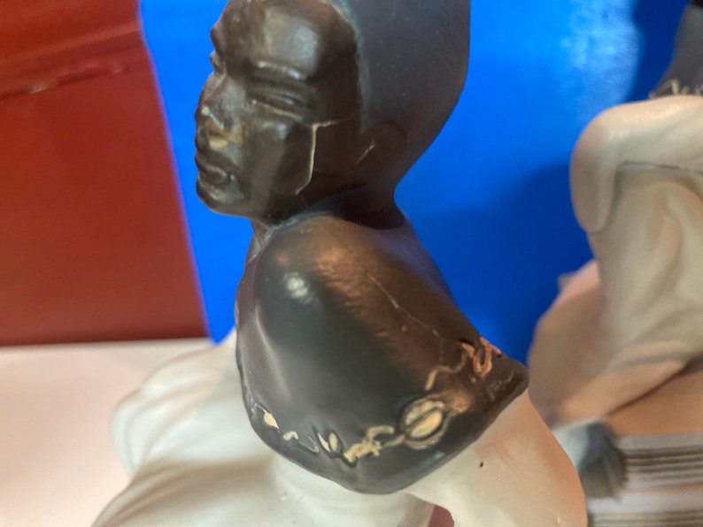 May include: A ceramic figurine featuring a black-skinned bust with a dark gray head and shoulders. The figurine has a white base and is set against a blue and red background. The bust is a side profile.