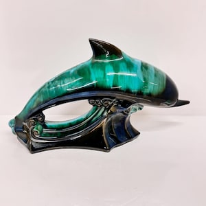 May include: A ceramic dolphin figurine with a green and black glaze. The dolphin is leaping out of the water, with its tail and fins outstretched. The base of the figurine is also decorated with a wave design.