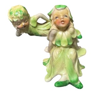 Könnte beinhalten: Eine Vintage-Porzellanfigur mit zwei Figuren. Eine Figur liegt, die andere sitzt. Beide Figuren tragen hellgrüne und cremefarbene Outfits mit blattartigen Details. Die sitzende Figur hat rosige Wangen und einen offenen Mund.