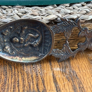 Puede incluir: Una cuchara de latón vintage con un mango decorativo y un diseño detallado de una figura en la parte delantera. La cuchara tiene un acabado deslustrado.