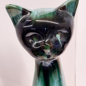 Puede incluir: Una figurita de gato de cerámica negra y verde con un acabado brillante. El gato tiene un cuerpo largo y delgado y una cabeza estilizada con ojos grandes en forma de almendra.
