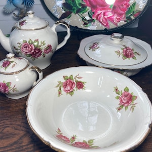 Puede incluir: Un juego de té vintage con un patrón de rosas. El juego incluye una tetera, un azucarero, una jarra de crema y un tazón grande para servir. Las piezas son blancas con ribetes dorados y presentan rosas rosadas.