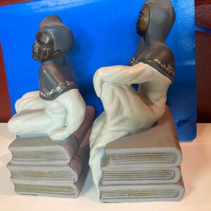 May include: Pair of ceramic bookends featuring figures in a seated pose atop stacks of books. The figures have dark skin, dark head coverings, and white robes. The books are gray with gold-colored edges.