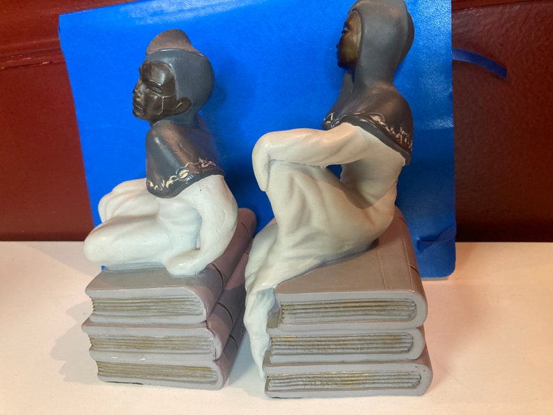 May include: Pair of ceramic bookends featuring figures in a seated pose atop stacks of books. The figures have dark skin, dark head coverings, and white robes. The books are gray with gold-colored edges.