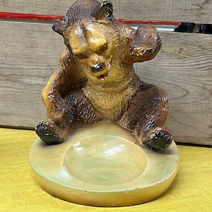 May include: A ceramic ashtray shaped like a bear sitting down. The bear is brown and has a light brown belly. The ashtray is a light brown color with a green center.