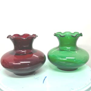 Puede incluir: Dos jarrones de cristal con bordes ondulados. Uno es de color rojo intenso y el otro verde vibrante. Ambos tienen un cuerpo redondeado y una parte superior ondulada y acampanada. Los jarrones son probablemente decorativos.