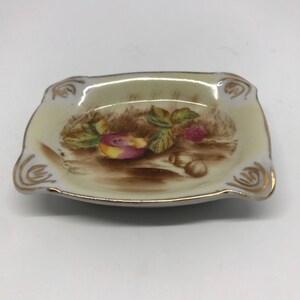 May include: A small, square, white ceramic ashtray with a gold rim. The ashtray has a painted design of fruit and leaves on a brown background.