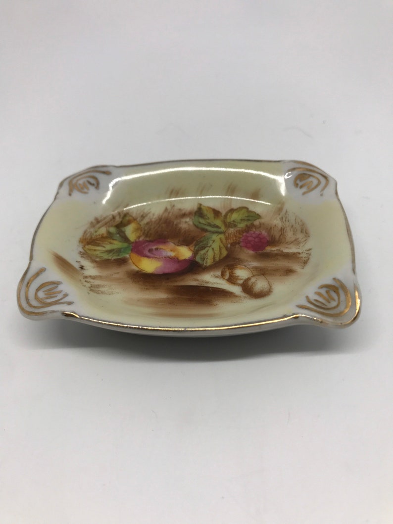 May include: A small, square, white ceramic ashtray with a gold rim. The ashtray has a painted design of fruit and leaves on a brown background.