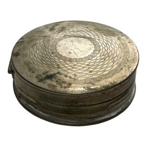 Könnte beinhalten: Kleine, runde, antike Metallbox in Silberoptik. Der Deckel weist ein geätztes, konzentrisches Wellenmuster um einen Mittelkreis auf. Die Box weist Alters- und Gebrauchsspuren auf, mit etwas Anlaufen und Verfärbungen, was auf Vintage hindeutet.