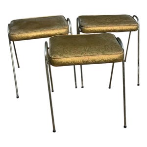 May include: Three vintage vanity stools with gold-colored, patterned cushions and chrome legs. The rectangular cushions have a floral design and are trimmed with a darker gold piping. The legs are slender and slightly angled.