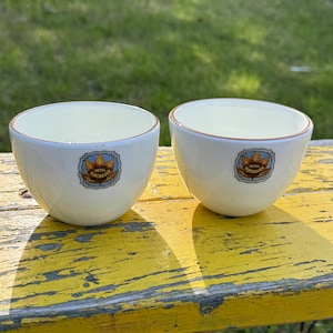 May include: Two white ceramic cups with a gold rim and a blue and gold logo that says "Hotel Vancouver" on a yellow wooden surface.