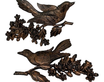 Pode incluir: Duas esculturas de pássaros em bronze pousadas em galhos floridos. Os pássaros têm penas e bicos detalhados, e os galhos apresentam flores e folhas delicadas. Decoração de parede.
