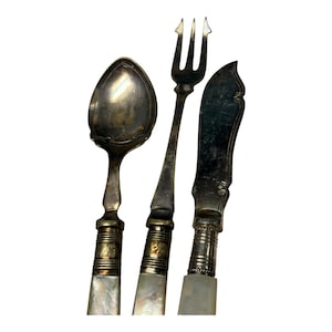 Op de afbeelding: Een set antiek zilverwerk, waaronder een lepel, vork en mes. De lepel heeft een ovale kom met decoratieve randen. De vork heeft drie tanden. Het mes heeft een gebogen lemmet. Elk stuk heeft een parelmoeren handvat en sierlijke metalen accenten.