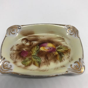 May include: A small, square, cream-colored ceramic ashtray with a gold rim. The ashtray features a brown and green floral design with a pink and yellow fruit.