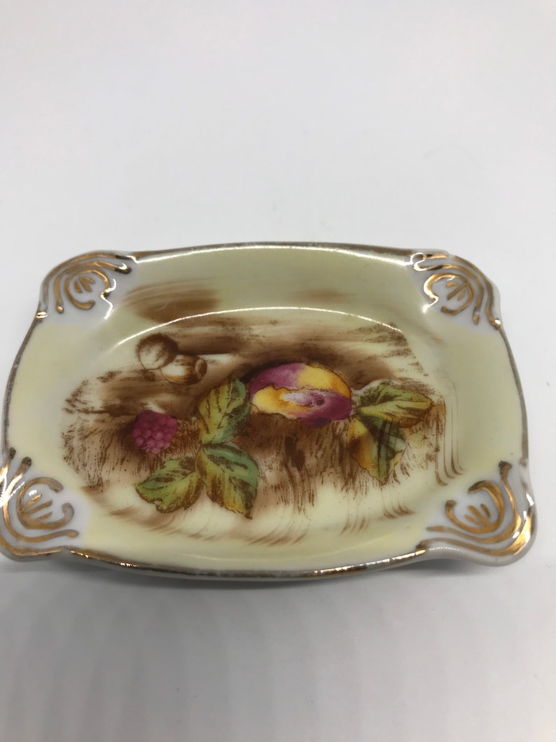 May include: A small, square, cream-colored ceramic ashtray with a gold rim. The ashtray features a brown and green floral design with a pink and yellow fruit.