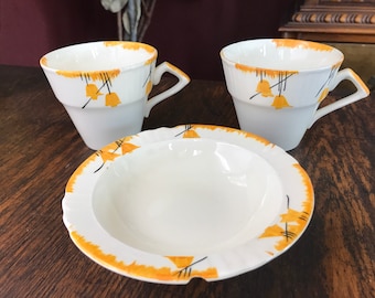Pode incluir: Conjunto de chá branco com um design decorativo de folhas laranja e pretas. O conjunto inclui duas chávenas e um pires. As chávenas têm um design de pega único. O conjunto está numa superfície de madeira.