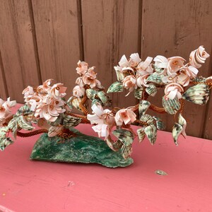 Puede incluir: Un árbol decorativo hecho de conchas marinas, con flores rosas y blancas y hojas verdes, sobre una base de piedra verde.
