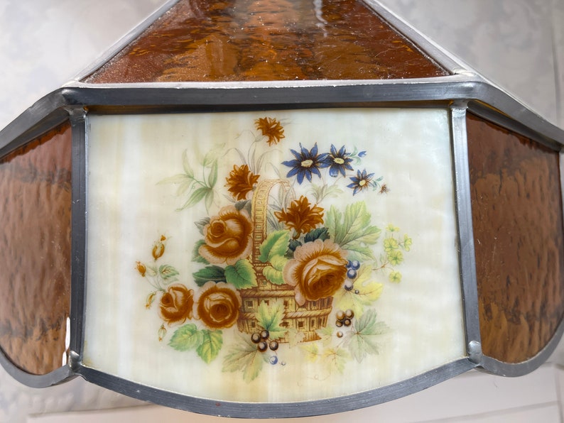 Puede incluir: Una pantalla de l&aacute;mpara de vidrio tintado con un dise&ntilde;o floral. La pantalla est&aacute; hecha de vidrio transparente con un borde marr&oacute;n y un centro blanco. El dise&ntilde;o floral presenta una cesta de flores, incluyendo rosas, margaritas y bayas.