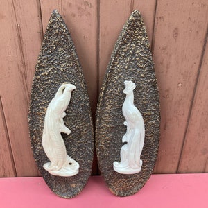 May include: Two wall plaques featuring white ceramic birds mounted on dark brown, textured, teardrop-shaped backings. Each plaque displays a glossy, off-white bird figure in profile, set against a rough, dark brown surface with gold highlights.