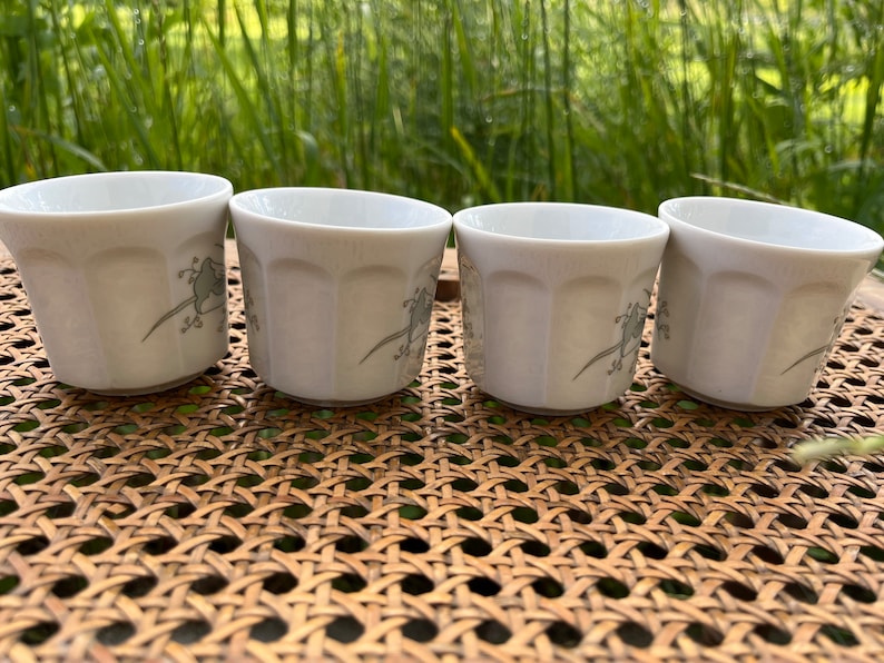 May include: Four white ceramic cups with a floral design. The cups are sitting on a woven wicker surface.