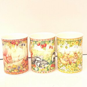 May include: Three ceramic mugs with woodland animal designs. Each mug features a different scene: squirrels, badgers, and rabbits. The mugs have a cream-colored background with colorful illustrations of nature scenes, berries, and flowers. The rims are decorated with a red and yellow border.