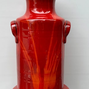 Peut inclure: Un vase en céramique rouge avec deux anses et un motif orange stylisé. Le vase a une finition brillante.