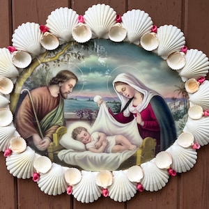 May include: A white seashell frame with pink accents surrounds a painting of the Holy Family. The painting depicts Mary, Joseph, and the baby Jesus in a manger.