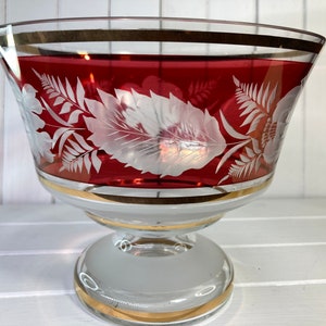 May include: A red glass bowl with a gold rim and white etched floral design. The bowl sits on a clear glass pedestal with a gold rim.