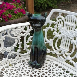 Può includere: Una statuetta di gatto in ceramica con una smaltatura lucida che sfuma dal nero profondo alla base al verde smeraldo brillante sulla parte superiore. Il gatto è raffigurato in posizione seduta con orecchie a punta, occhi a mandorla e finitura liscia.