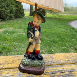 May include: A vintage lamp base featuring a figurine of a boy wearing a green jacket, brown shorts, and a red scarf. The boy is holding a walking stick and is standing on a green base with brown accents.