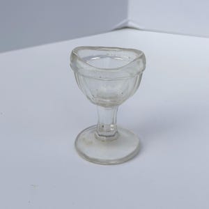 May include: A clear glass eye wash cup featuring a contoured rim designed to fit around the eye socket, a short stem, and a wide circular base. The cup has a smooth, transparent finish with subtle vertical ribbing along the exterior bowl.