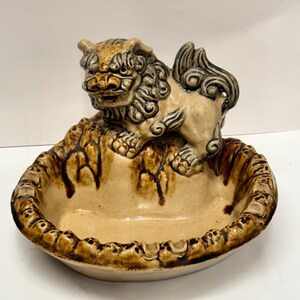 May include: A ceramic ashtray with a brown and beige glaze. A small, detailed ceramic lion figurine sits on top of the ashtray.