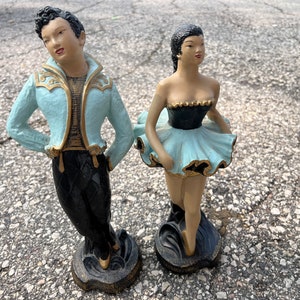 Peut inclure: Une paire de figurines en céramique vintage représentant un danseur et une danseuse. La figurine masculine porte une veste bleue et dorée avec un pantalon noir. La figurine féminine porte un tutu bleu et noir. Les deux figurines sont debout sur une base noire.