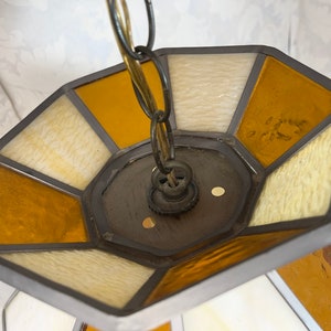 Puede incluir: Una l&aacute;mpara de techo de cristal de colores con una cadena colgando de ella. La l&aacute;mpara es octogonal y tiene un marco de metal. Los paneles de vidrio son en tonos de amarillo, blanco y marr&oacute;n.