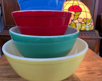 May include: A stack of four vintage mixing bowls in various colors: blue, red, green, and yellow. The bowls are nested inside each other, showcasing their different sizes and hues. The bowls are made of a glossy material.