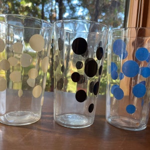 Puede incluir: Tres vasos de vidrio transparente para beber con lunares de diferentes colores. El primer vaso tiene lunares blancos, el segundo tiene lunares negros y el tercero tiene lunares azules.