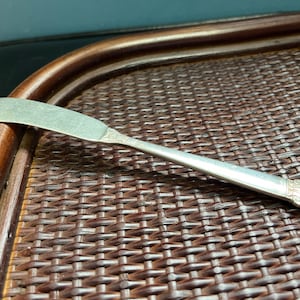 Puede incluir: Un cuchillo de mantequilla de plata con un mango adornado, descansando sobre una superficie de mimbre marrón.