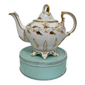 May include: White porcelain teapot with gold detailing and floral patterns. The teapot features a curved spout, a loop handle, and a lid with a gold finial. It rests on a light blue tin.