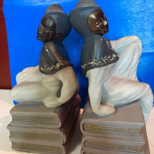 May include: Pair of ceramic bookends featuring figures in a seated pose atop stacks of books. The figures have black faces and wear dark gray head coverings. The books are gray with gold-colored edges, and the figures are dressed in white.