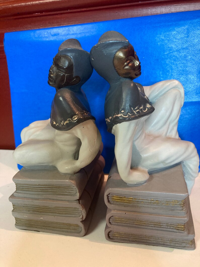 May include: Pair of ceramic bookends featuring figures in a seated pose atop stacks of books. The figures have black faces and wear dark gray head coverings. The books are gray with gold-colored edges, and the figures are dressed in white.