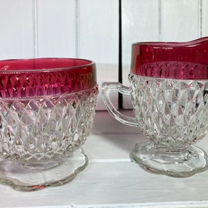 Puede incluir: Un juego vintage de azucarero y jarra de crema de vidrio con un patrón de diamante transparente y un borde rojo rubí.