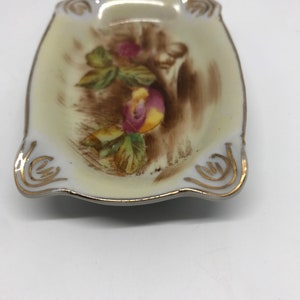May include: A small, white porcelain dish with a gold rim and a floral design. The dish has a scalloped edge and is perfect for holding small items.