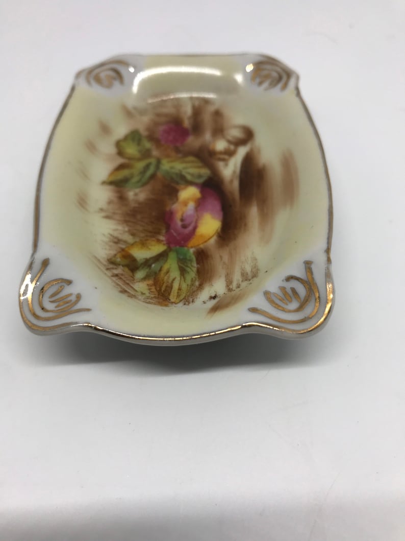 May include: A small, white porcelain dish with a gold rim and a floral design. The dish has a scalloped edge and is perfect for holding small items.