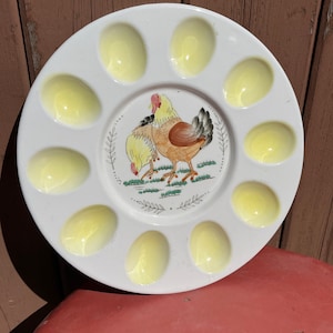Op de afbeelding: Witte keramische eierplaat met 10 gele, eivormige inkepingen. De plaat heeft een geschilderd ontwerp van twee kippen in een grasveld.