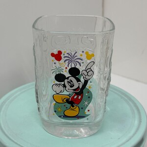 Puede incluir: Vaso de vidrio transparente con un diseño de Mickey Mouse. El diseño presenta a Mickey en una pose festiva con detalles rojos, amarillos y negros, además de confeti y fuegos artificiales. El vaso tiene un patrón texturizado en los lados.