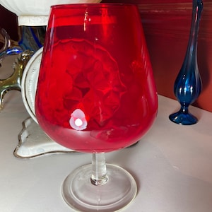 Może przedstawiać: Duży, przezroczysty kielich szklany z czerwonym, wirującym płynem w środku. Kielich ma szeroką, okrągłą czaszę i wysoką, smukłą nóżkę.