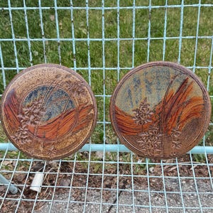 May include: Two circular ceramic plates with a rustic design. Each plate features a brown, orange, and blue colour scheme with floral and abstract patterns. The plates are displayed on a light blue metal grid.