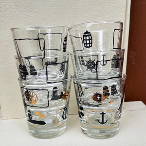 May include: A set of six clear glass shot glasses. Each glass is decorated with black and gold nautical designs, such as anchors, lighthouses, and ship wheels. The glasses are stacked in two rows of three.
