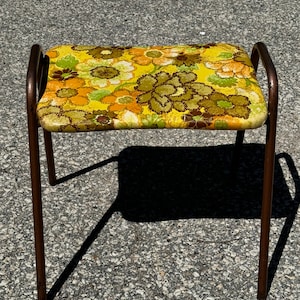 Puede incluir: Taburete vintage de metal con asiento rectangular. El asiento presenta un estampado floral en tonos amarillos, verdes y marrones. El marco de metal es de color marrón oscuro. Dimensiones: aproximadamente 40 cm de largo.