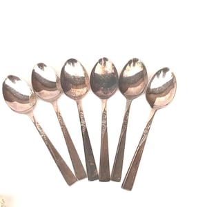 May include: Six vintage silver spoons arranged on a white background. The spoons have oval bowls and ornate, engraved handles. The spoons are made of a shiny, reflective metal, likely silver or a silver-plated alloy. The spoons are a classic style.